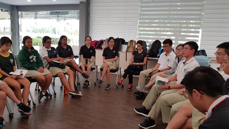 Students and adults sit in a circle on chairs in a bright room, appearing to hold a group discussion.